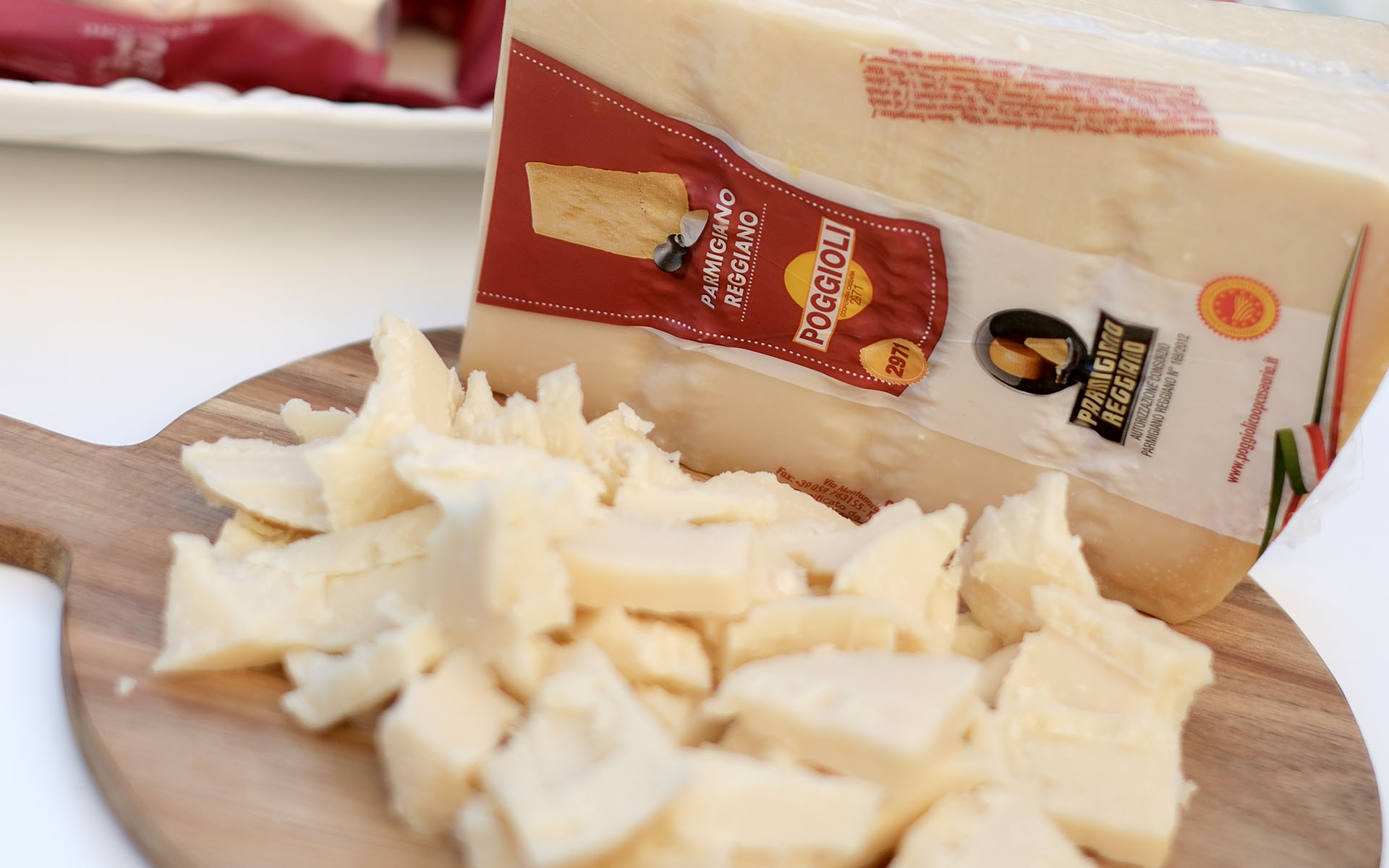 L'immagine mostra un tagliere di legno con sopra una quantità di Parmigiano Reggiano a pezzi. I pezzi di formaggio sono di diverse dimensioni e forme, con superfici irregolari tipiche del formaggio spezzato a mano, mostrando una consistenza friabile e granulosa. Accanto al formaggio sbriciolato, su un lato del tagliere, si trova un pezzo più grande di Parmigiano Reggiano confezionato. La confezione è parzialmente aperta e mostra una sezione di etichetta rossa con la scritta "Parmigiano Reggiano Poggioli" e un logo che evidenzia l'autenticità del prodotto. Il colore del formaggio è un giallo pallido, tipico del Parmigiano Reggiano stagionato. Sullo sfondo si intravede un piatto bianco, decorato con un tovagliolo rosso scuro. L'immagine è ben illuminata, enfatizzando la texture e il colore del formaggio.