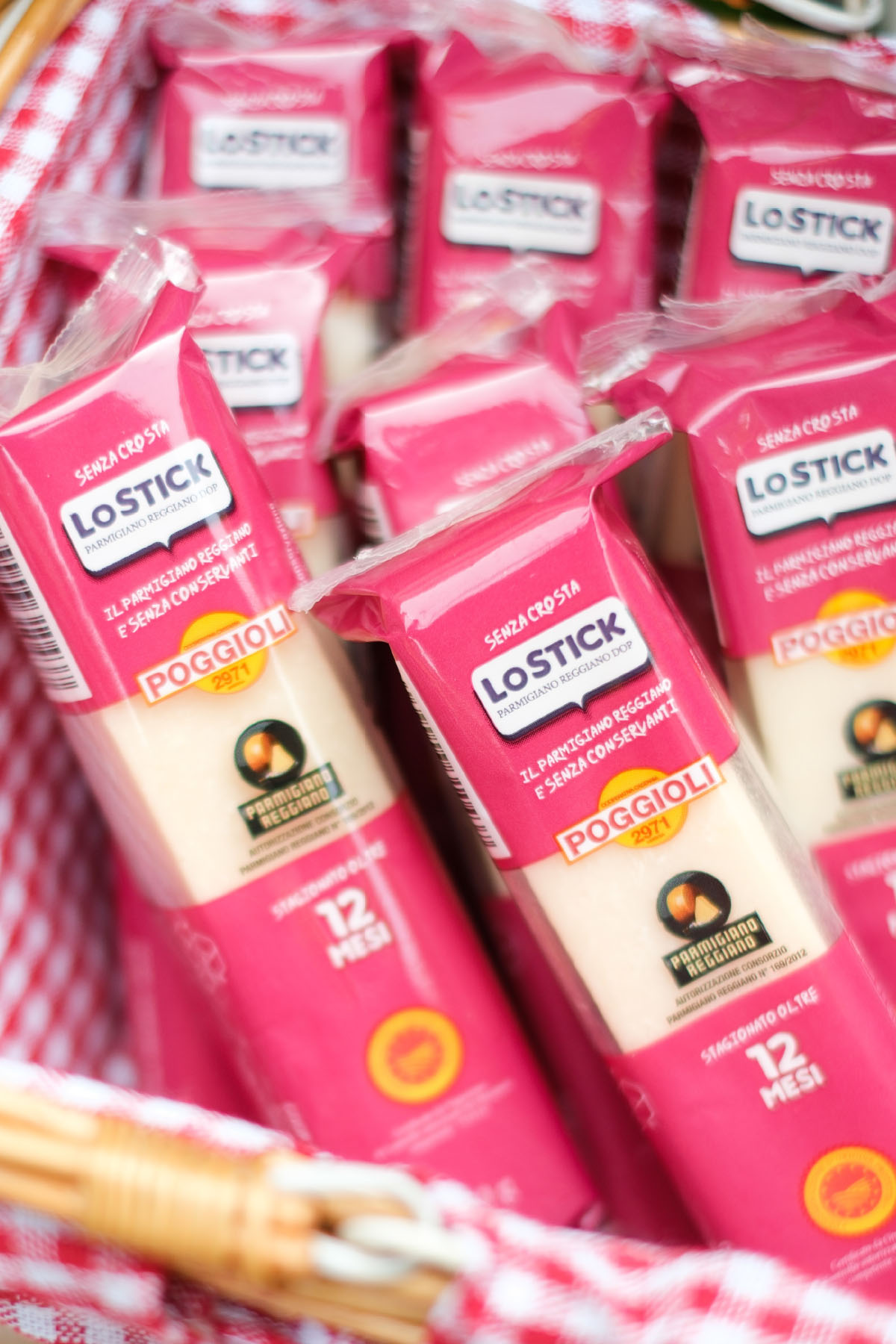 L'immagine mostra un cesto da picnic in vimini, rivestito all'interno con un tessuto a quadri bianchi e rossi. Nel cesto sono disposte numerose confezioni di plastica rosa contenenti "Lo Stick Parmigiano Reggiano DOP".
Sul fronte di ogni confezione, in alto, c'è la scritta bianca "SENZA CROSTA". Sotto, su fondo bianco, si legge "LO STICK Parmigiano Reggiano DOP". Più in basso, sempre in bianco, è riportata la dicitura "Il Parmigiano Reggiano è senza conservanti".
Al centro della confezione è presente un'etichetta circolare con il logo "Poggioli 2971" in rosso e oro. Sotto il logo, si trova il marchio circolare nero e oro del "Parmigiano Reggiano", indicandone l'autenticità. La scritta in bianco "stagionato oltre 12 mesi" descrive il tempo di stagionatura del formaggio.
Il bordo superiore delle confezioni presenta un’apertura trasparente che permette di vedere il contenuto. L'illuminazione naturale del sole fa risaltare i colori rosa e bianco delle confezioni e dell'interno del cesto, creando un'atmosfera invitante e conviviale, ideale per un pranzo all'aperto o una merenda.