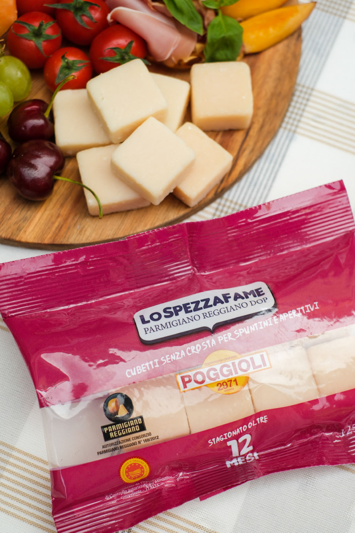 L'immagine presenta una scena appetitosa e ben curata. Al centro, su un piano di legno, sono disposti cubetti di Parmigiano Reggiano senza crosta. Intorno ai cubetti si vedono vari ingredienti freschi: pomodorini rossi con piccoli gambi verdi, ciliegie scure e lucide, uva verde, un peperone giallo tagliato a fette, fette di prosciutto arrotolate e foglie di basilico verde.
In primo piano, c'è una confezione di plastica di colore rosa scuro con la scritta "Lo Spezzafame Parmigiano Reggiano DOP", accompagnata dalla didascalia "Cubetti senza crosta per spuntini e aperitivi".
Il marchio "POGGIOLI 2971" è ben visibile, insieme al logo del Parmigiano Reggiano che ne conferma l'autenticità. Si nota la dicitura "stagionato oltre 12 mesi" in bianco sulla confezione. Il tutto è posto su una tovaglia bianca con quadri sottili grigio chiaro, che dona un aspetto elegante e semplice alla scena.