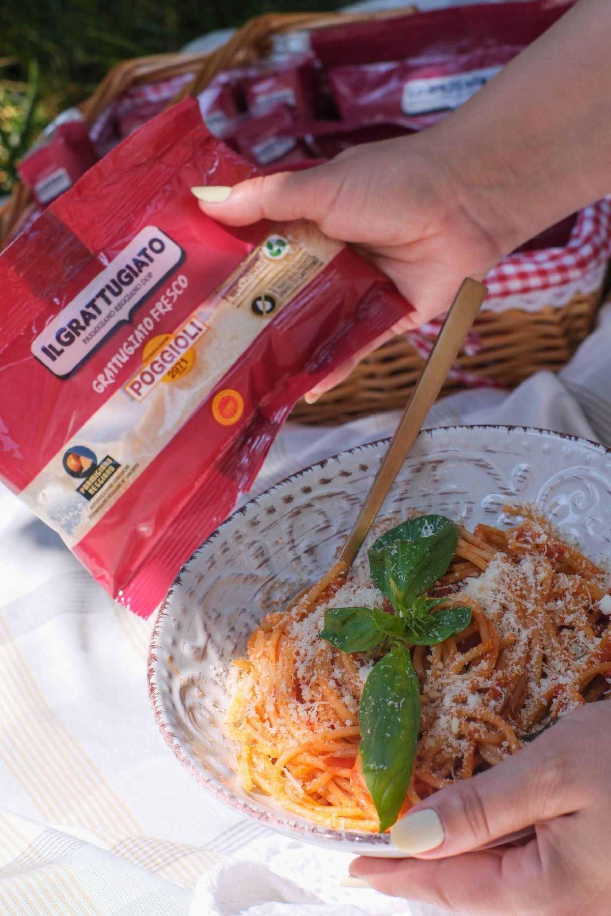 Sul piatto di ceramica bianca decorata, sono serviti spaghetti conditi con salsa di pomodoro. Gli spaghetti sono coperti da una spolverata di formaggio grattugiato e adornati con foglie di basilico fresco, che aggiungono un tocco di verde brillante. La forchetta dorata è infilata nella pasta, pronta per essere usata.
La mano della persona, con unghie dipinte di un colore chiaro, sostiene il piatto con delicatezza. Nell'altra mano, tiene una confezione di plastica di colore rosso vivo con dettagli lucidi.
Sulla confezione si leggono diverse scritte: "IL GRATTUGIATO" in bianco con bordo blu, "GRATTUGIATO FRESCO" e "POGGIOLI 2971" in bianco su sfondo rosso. Ci sono anche simboli che indicano informazioni nutrizionali e certificazioni, tra cui il marchio del Parmigiano Reggiano.
In secondo piano, sfocato, si intravede un cesto di vimini con altre confezioni simili, suggerendo una scena di picnic. Il cesto è foderato con un panno a quadretti bianchi e rossi, e l'ambiente circostante è pieno di verde, indicando che si tratta di una giornata all’aperto.
Questa immagine trasmette un senso di gusto e freschezza, combinando il piacere del cibo italiano con la semplicità di un pasto goduto nella natura.