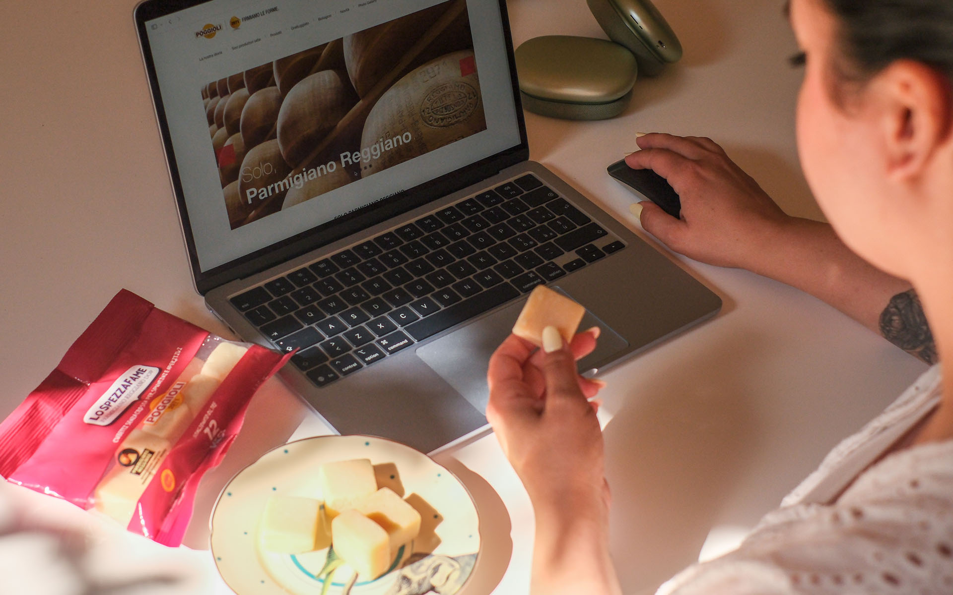 L'immagine mostra una persona seduta a un tavolo bianco, impegnata al computer portatile, e accanto a essa ci sono degli snack di Parmigiano Reggiano.
Davanti a sé, la persona ha un laptop aperto che mostra sullo schermo una pagina web dedicata al Parmigiano Reggiano, con l'immagine di grandi forme di formaggio e il logo "Poggioli". La mano destra della persona è poggiata su un mouse nero, mentre con la sinistra tiene un cubetto di Parmigiano Reggiano vicino alla bocca, pronta a gustarlo. Le unghie sono curate e dipinte di un colore chiaro.
Sul tavolo, sulla sinistra, si vede una confezione di plastica rosso scuro, con l'etichetta "Lo Spezzafame" e il logo "Poggioli 2971". La confezione è aperta e alcuni dei cubetti sono stati sistemati su un piccolo piatto di ceramica decorato con un bordo verde e piccoli motivi blu, anch'esso sul tavolo.
In alto a destra dell'immagine, si notano due custodie tondeggianti di colore verde chiaro. La luce nell'ambiente è calda, creando un’atmosfera accogliente e rilassante.