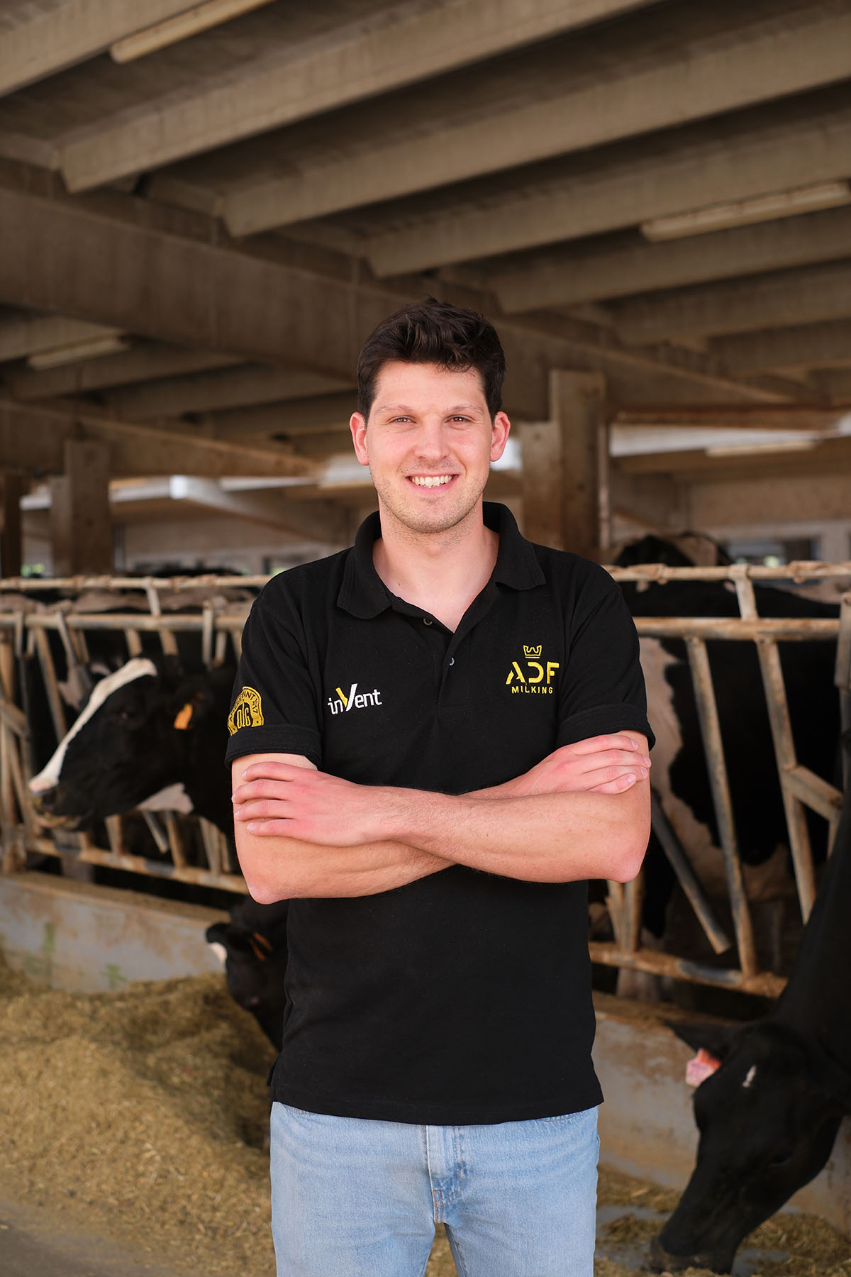 L'immagine mostra un uomo sorridente all'interno di una stalla. Indossa una polo nera con loghi gialli sulla parte sinistra del petto e sulla manica destra, e un paio di jeans chiari. L'uomo ha le braccia incrociate sul petto e si trova davanti a una fila di mucche che si sporgono attraverso una struttura di metallo per mangiare il fieno distribuito sul pavimento.
La stalla ha un soffitto in cemento con travi visibili e grandi aperture che lasciano entrare la luce. L’ambiente appare pulito e ordinato. Le mucche, di colore nero con macchie bianche, sono in una fila ordinata dietro la barriera metallica. Sullo sfondo, la luce naturale illumina ulteriormente la scena, creando un'atmosfera chiara e accogliente. L'intera immagine evoca un senso di tranquillità e dedizione al lavoro con gli animali.