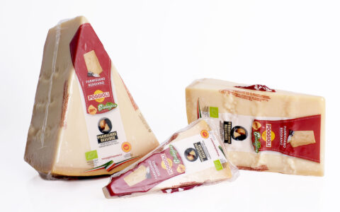 L’immagine mostra tre grandi pezzi di formaggio Parmigiano Reggiano “Poggioli 2971 Biologico”, disposti in primo piano su una superficie bianca, davanti a uno sfondo anch’esso perfettamente bianco che accentua la luminosità della scena. Tutti i pezzi sono confezionati e sigillati nella plastica trasparente, con etichette evidenti nella parte anteriore. Descrizione dei pezzi (da sinistra a destra): 1. A sinistra: - Un grande spicchio triangolare di Parmigiano Reggiano, alto circa trenta centimetri e largo alla base. - La crosta marrone chiaro è visibile sul lato inferiore, mentre la parte superiore e le facce laterali mostrano la pasta chiara, di colore avorio, compatta e leggermente granulosa. - Sulla faccia principale, vi è una grande etichetta rettangolare verticale. La parte superiore dell’etichetta è di un rosso-bordeaux lucido; vi è illustrato un piccolo triangolino di formaggio. Subito sotto sono riportate le scritte “PARMIGIANO REGGIANO”, “POGGIOLI 2971”, e la parola “Biologico” in verde con grafia vivace, simile a una scrittura a mano. - Al centro della stessa etichetta, compare il logo “Parmigiano Reggiano” tondo, nero e oro, con l’immagine di una forma di formaggio tagliata a metà, segno di originalità e autenticità del prodotto. - In basso a sinistra la bandierina italiana, in basso a destra una piccola etichetta circolare rossa con il simbolo DOP. 2. Al centro in basso: - Un altro spicchio di Parmigiano Reggiano, più piccolo e sottile, è appoggiato leggermente sovrapposto agli altri pezzi. - Ha la stessa confezione: parte superiore dell’etichetta color rosso scuro, scritta “POGGIOLI 2971 Biologico” in grande, e il logo del Parmigiano Reggiano. Si nota inoltre una targhetta piccola e verde che indica la certificazione di prodotto biologico. - La confezione è completamente trasparente lungo il bordo, lasciando intravedere le irregolarità della pasta compatta del formaggio. 3. A destra: - Un grande parallelepipedo rettangolare di Parmigiano Reggiano, più largo e basso rispetto agli altri pezzi. - Anche qui la crosta color beige scuro corre lungo la base, mentre la parte di pasta mostra tipiche venature bianche dovute alla stagionatura. - L’etichetta è simile alle precedenti: parte superiore rosso scuro bordeaux, il logo Poggioli 2971 in giallo, la scritta “Parmigiano Reggiano” e la dicitura “Biologico”. La confezione trasparente enfatizza l’omogeneità della pasta e la texture leggermente ruvida e asciutta del formaggio. Ulteriori dettagli: - Tutte le etichette riportano chiaramente simboli e certificazioni di qualità, come la sigla DOP e il marchio europeo verde di prodotto biologico (“leaf mark” con foglia stilizzata). - Nessun altro oggetto è presente nella scena, che si concentra esclusivamente sui tre formaggi. - La superficie bianca riflette leggermente le basi dei pezzi di formaggio, conferendo un senso di pulizia, freschezza e ordine. Sintesi sensoriale: L’immagine comunica qualità e artigianalità. La presentazione limpida e le etichette colorate risaltano sul fondo bianco, riflettendo la purezza e la genuinità del Parmigiano Reggiano. La forma geometrica dei pezzi e l’irregolarità della superficie suggeriscono un prodotto autentico, stagionato a lungo e pronto per essere assaporato, sia a tavola che come ingrediente in cucina.