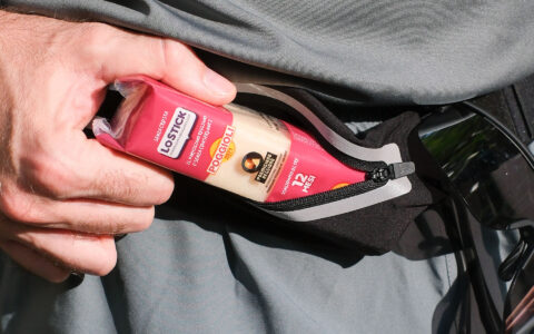 L’immagine è un primo piano molto dettagliato di una mano maschile che estrae un piccolo snack confezionato da una tasca con cerniera di un capo d’abbigliamento sportivo, probabilmente dei pantaloncini o una giacca tecnica di colore grigio. La mano è leggermente aperta e afferra lateralmente la confezione, lasciando ben visibili le nocche e le vene della mano, che è di carnagione chiara e mostra una leggera peluria. Il protagonista dell’immagine è uno snack confezionato: si tratta di un bastoncino di formaggio Parmigiano Reggiano. La confezione, di forma allungata e rettangolare, è di plastica e ha colori prevalentemente rosso scuro in alto e crema sul fondo, con scritte bianche, nere e arancioni. Sulla superficie della confezione si leggono varie informazioni: il marchio “LOSTICK”, ben visibile su uno sfondo bianco, accompagnato dalla scritta in piccolo “IL PRATICO PARMIGIANO REGGIANO” e “SENZA CONSERVANTI”. Subito sotto c’è il logo dell’azienda “POGGIOLI 2971”. In un riquadro nero è riportato il simbolo del Parmigiano Reggiano con la scritta “Parmigiano Reggiano”, seguita in verticale dalla dicitura “stagionatura oltre 12 mesi”. La tasca da cui viene estratto lo snack è nera, dotata di una cerniera nera e un bordo grigio chiaro, e sembra far parte di un indumento sportivo. Accanto, in basso a destra nell’immagine, si intravedono degli occhiali sportivi da sole neri, probabilmente appesi al bordo dell’indumento. Lo sfondo appare completamente occupato dal tessuto grigio dei pantaloni o della giacca, leggermente piegato e increspato. La luce naturale illumina la scena da sinistra, creando ombre decise sulle pieghe del tessuto e sulla mano. Nel complesso, l’immagine comunica un senso di praticità e modernità, sottolineando la portabilità di uno snack salato di alta qualità, facilmente trasportabile durante attività sportive o all’aperto. L’aspetto generale è pulito, ordinato e nitido, con particolare attenzione ai dettagli della mano, della confezione e della tasca sportiva.