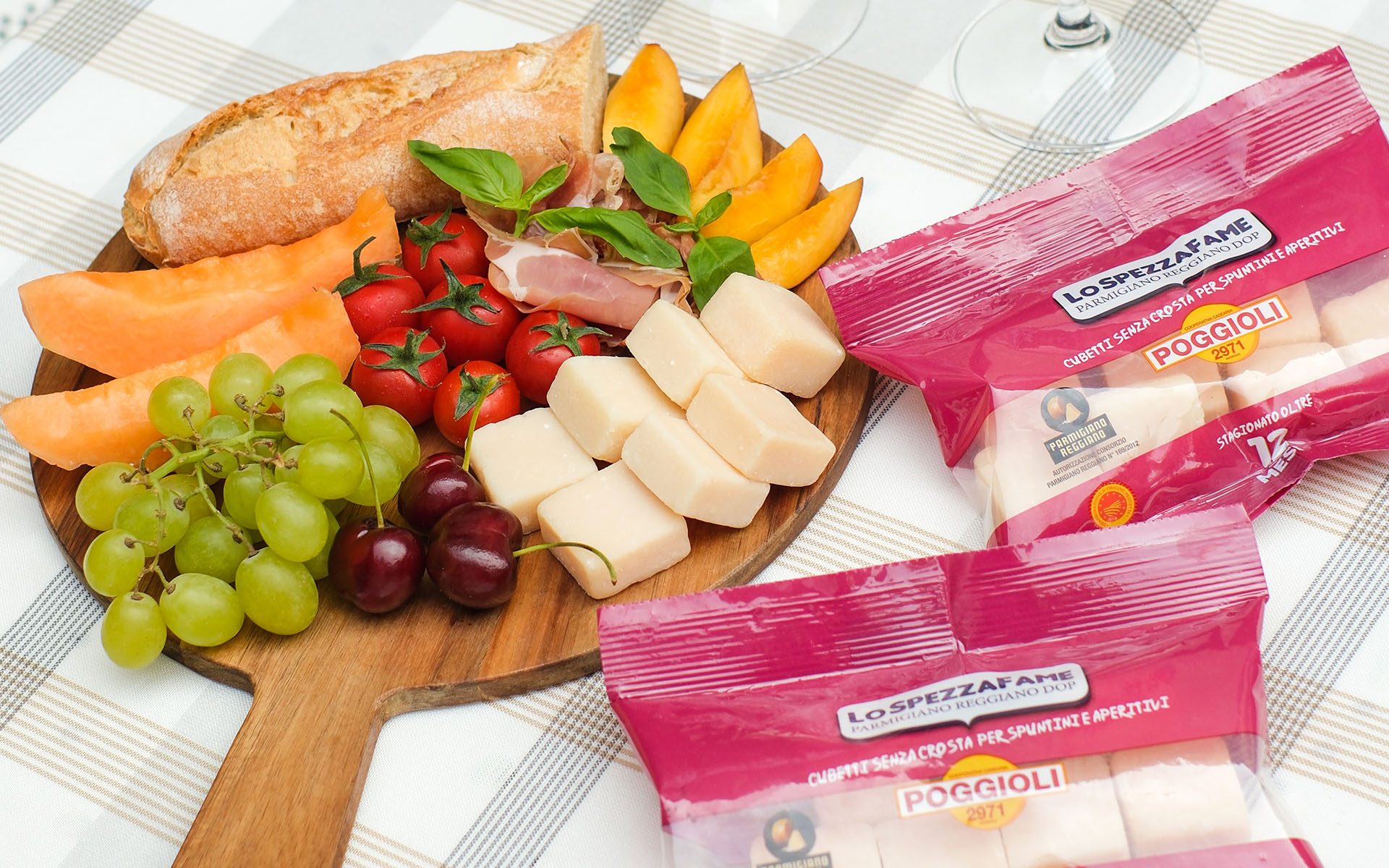 L’immagine mostra una scena conviviale tipica di un picnic estivo o di un pranzo leggero all’aperto, ricca di colori e dettagli. Tutti gli elementi poggiano su una tovaglia a quadrettoni chiari nei toni del bianco, beige e grigio chiaro. Al centro dell’immagine si trova un grande tagliere di legno rotondo con manico. Su questo tagliere sono disposti ordinatamente diversi alimenti freschi: - In alto, un filone di pane rustico dalla crosta dorata e spessa. - Sul lato sinistro, quattro fette di melone arancione brillante, morbide e succose. - Accanto, un bel grappolo di uva verde, i chicchi sono tondeggianti e traslucidi, perfetti e lucidi. Poco sotto ci sono due ciliegie di colore violaceo scuro e brillante, con picciolo. - Più verso il centro, si trovano diversi pomodori ciliegini, piccoli, rotondi e di un rosso vivace, ancora attaccati al rametto verde. - Sopra i pomodori, al centro del tagliere, sono adagiate alcune fette di prosciutto crudo leggero, piegate morbidamente e decorate con foglie di basilico fresche e dal colore verde intenso. - Sul lato destro del tagliere sono disposte sei fette di pesca matura, di un arancio dorato intenso, con la buccia rosata. - Vicino alle pesche, in basso a destra sul tagliere, sono ordinati dei cubetti grandi di Parmigiano Reggiano Dop, di colore panna, un po’ opachi e con superficie leggermente irregolare. Sulla destra dell’immagine, al di fuori del tagliere, si trovano due confezioni di Parmigiano Reggiano Dop a cubetti, marchio "LO SPEZZAFAME" Poggioli 2971, con confezione di colore fucsia trasparente che lascia intravedere i cubetti di formaggio all’interno. Sulle confezioni si possono leggere le diciture: "Cubetti senza crosta per spuntini e aperitivi", "Stagionato oltre 12 mesi", e i loghi di certificazione e marchio Parmigiano Reggiano. Nell’angolo alto, sullo sfondo, sono visibili parte dei calici da vino trasparenti poggiati sulla tovaglia, suggerendo un’atmosfera di relax e convivialità, come durante un aperitivo o una merenda tra amici. Sintesi sensoriale: L’immagine comunica freschezza, genuinità e varietà tipiche di un pranzo all’italiana: i toni caldi di melone, pesche e pomodori si mescolano al verde delle uve e del basilico, mentre i cubetti di Parmigiano e le fette di pane invitano ad assaporare consistenze differenti. Il profumo del basilico, la dolcezza della frutta fresca, il sapore intenso del formaggio e la fragranza rustica del pane evocano un’esperienza gustativa ricca e autentica. L’ambiente trasmette armonia, accoglienza e il piacere della buona compagnia.