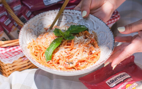 L’immagine mostra una scena luminosa e ravvicinata, centrata su un piatto di spaghetti all’italiana, tipicamente estiva e conviviale. In primo piano si vede un grande piatto rotondo di porcellana bianca, decorato con motivi in rilievo dal profilo leggermente frastagliato. Il piatto è tenuto da due mani: una mano lo regge lateralmente mentre l’altra, da sinistra, impugna un cucchiaio dorato con cui si mescola o si aggiunge del formaggio grattugiato sulla pasta. Gli spaghetti sono ben conditi con sugo di pomodoro rosso brillante e ricoperti generosamente da uno strato di formaggio Parmigiano Reggiano grattugiato fine, di colore chiaro, quasi bianco-crema, che crea una texture soffice sulla superficie del piatto. Guadagna il centro della scena una grande foglia di basilico verde, fresca, ancora umida, stesa orizzontalmente sopra la pasta e un piccolo ciuffo di basilico disposto più verticalmente affianco, donando al piatto un tocco di colore vivace e profumato. Sul lato sinistro dell’immagine si intravede, parzialmente fuori fuoco, un cesto da picnic in vimini decorato all’interno con una tovaglietta a quadretti rossi e bianchi. Dal cestino spuntano alcune confezioni di Parmigiano grattugiato “IL GRATTUGIATO”, riconoscibili dalla plastica lucida color bordeaux con una striscia bianca e le scritte nere. Una di queste confezioni è visibile anche nell’angolo in basso a destra dell’immagine, posata sulla tovaglia colorata a quadri beige, bianchi e grigi: riporta la dicitura “IL GRATTUGIATO PARMIGIANO REGGIANO POGGIOLI”. La luce della scena è naturale e calda, tipica di un pranzo all’aria aperta. Illumina il piatto, il formaggio e le mani con una luce diretta, mettendo in risalto le texture del formaggio, la lucentezza del basilico e la delicatezza dell’impasto degli spaghetti. La mano che regge il piatto ha unghie curate, smaltate di colore chiaro, dando un senso di cura e attenzione ai dettagli. Lo sfondo, pur sfocato, suggerisce un momento di relax su un prato, forse durante un picnic o una colazione sull’erba. Tra i dettagli secondari si notano lembi di tovaglia, fili d’erba e il bordo del cestino. L’atmosfera generale dell’immagine trasmette calore, appetito e il piacere della buona cucina italiana condivisa in compagnia, con il Parmigiano Reggiano protagonista sulla tavola. Sintesi sensoriale: La scena comunica freschezza, semplicità e gusto: si percepisce l’aroma intenso del basilico, il sapore ricco degli spaghetti al pomodoro, la morbidezza del formaggio appena grattugiato e il clima rilassato di un pasto consumato all’aria aperta, in un contesto familiare e accogliente.