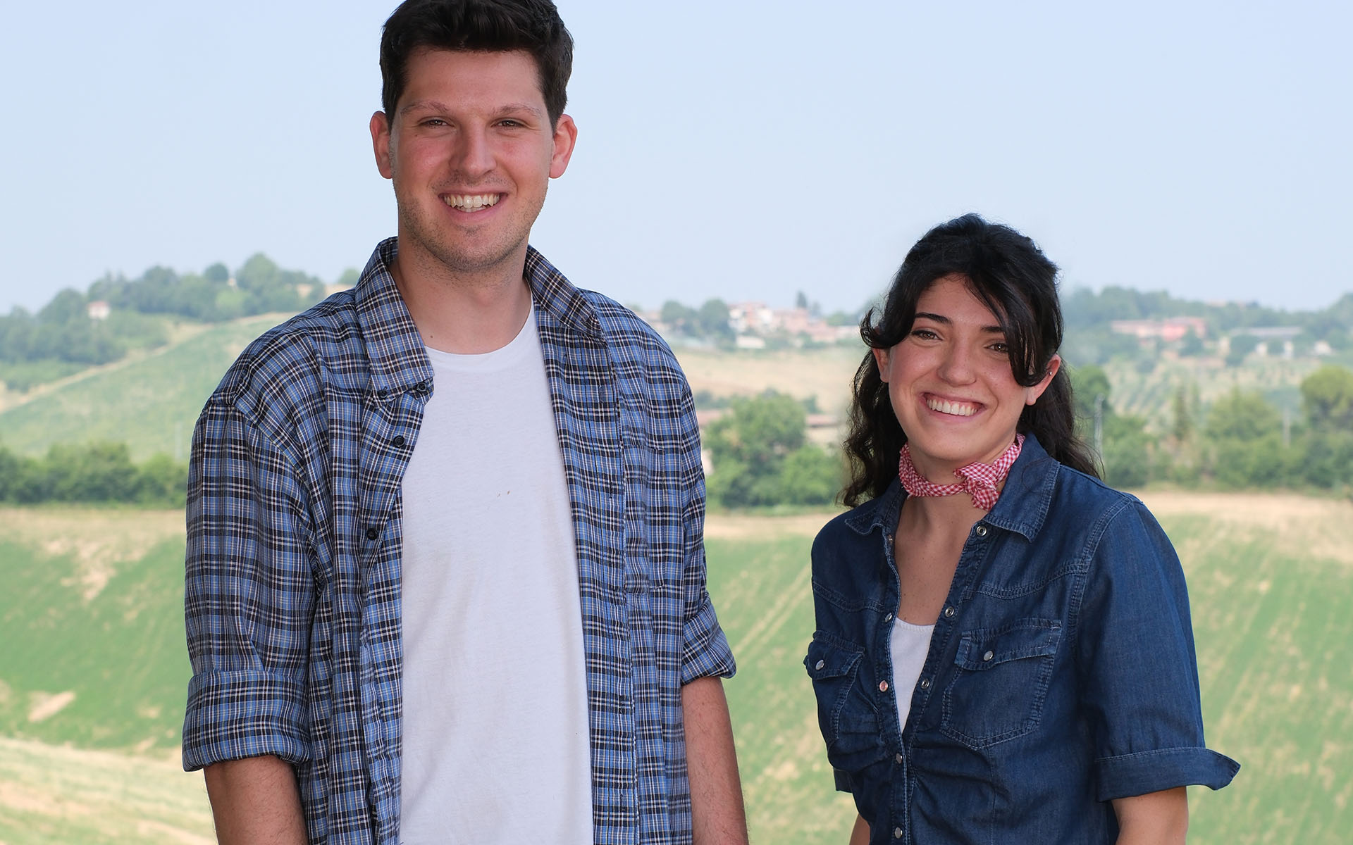 L’immagine mostra due giovani adulti in primo piano, posizionati uno accanto all’altro e leggermente rivolti verso la macchina fotografica. Sono entrambi sorridenti e trasmettono una sensazione di gioia e serenità. Sullo sfondo, si estende un ampio paesaggio collinare, caratterizzato da pendii verdi, campi coltivati e alcune macchie di alberi; il cielo è limpido e di un celeste molto chiaro, creando un’atmosfera luminosa e tipicamente rurale. La persona a sinistra nell’immagine è un uomo. Ha capelli corti, castani, e la carnagione chiara. Indossa una camicia a quadri azzurra e blu, con le maniche arrotolate fino ai gomiti. Sotto la camicia, porta una maglietta bianca. Ha un’espressione felice, sorride a denti visibili e guarda direttamente verso l’obiettivo. Alla sua destra c’è una donna, anche lei con la carnagione chiara e capelli scuri e lunghi, che indossa sciolti con una leggera frangia che le cade sulla fronte. Il suo abbigliamento consiste in una camicia di jeans blu, a maniche corte e con taschini sul petto, sotto la quale si intravede una magliettina bianca. Porta un foulard a scacchi rossi e bianchi annodato morbidamente intorno al collo, conferendole un aspetto fresco e informale. Anche lei sorride in modo aperto e simpatico. Lo sfondo è costituito da vaste colline di un verde intenso, dove si distinguono strisce di coltivazione e piccoli gruppi di alberi. In lontananza si nota un piccolo borgo con case sparse e qualche struttura agricola, immerso nel paesaggio rurale italiano. La giornata appare soleggiata, la scena molto luminosa e il paesaggio è nitido. Non sono presenti oggetti nelle mani delle due persone, né altri elementi che possano distogliere l’attenzione dai protagonisti o dal panorama agricolo alle loro spalle. L’abbigliamento dei due giovani e la naturalezza dei loro sorrisi suggeriscono che si trovano in un contesto campestre, probabilmente legato al lavoro o a una visita in ambiente rurale. In sintesi, l’immagine offre un ritratto positivo di due giovani immersi nella campagna, con lo sfondo di colline e paesaggi coltivati tipici della ruralità italiana. L’atmosfera generale è luminosa e serena, trasmettendo un senso di benessere e di connessione armoniosa con l’ambiente naturale circostante.