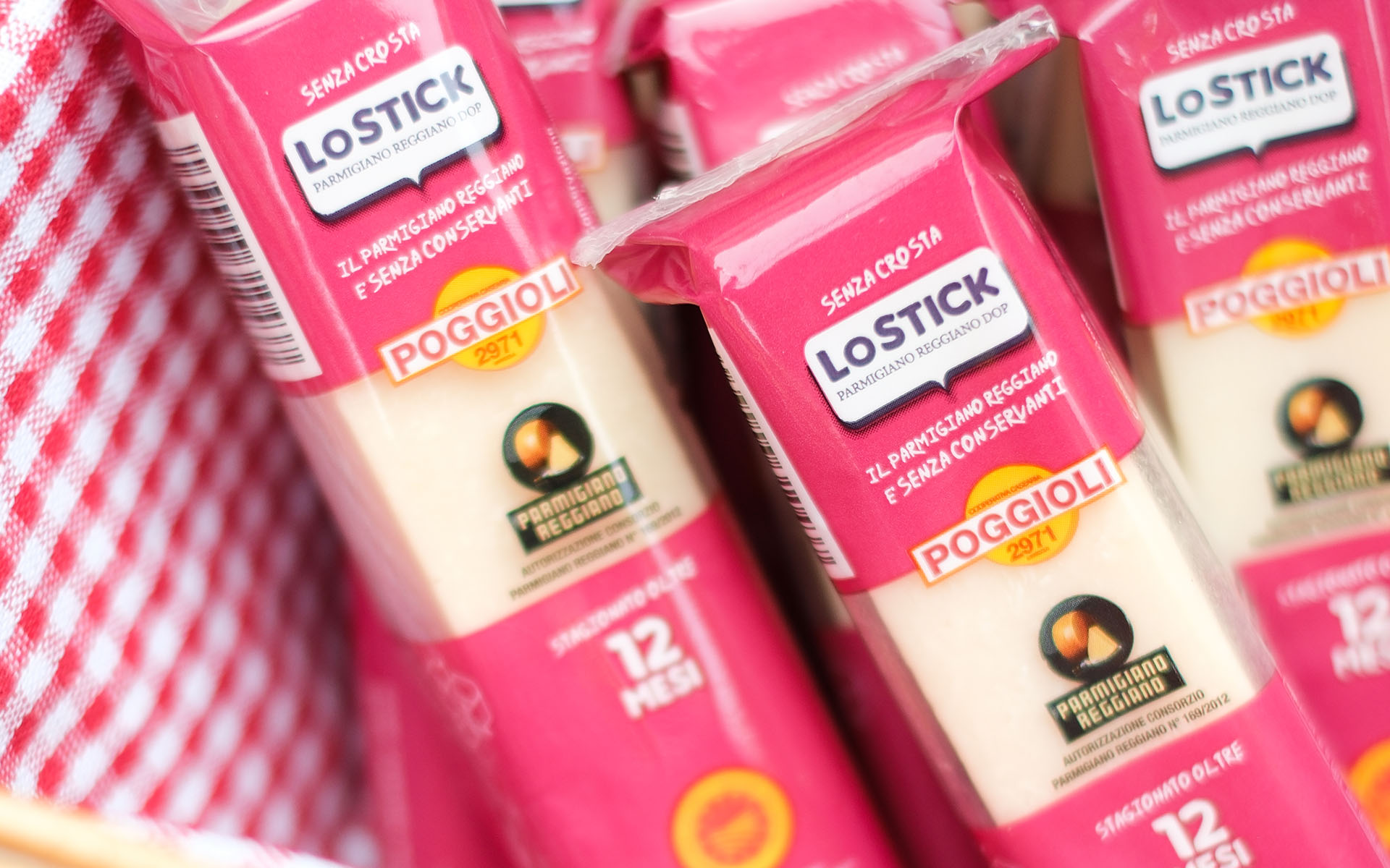 L’immagine mostra un primo piano ravvicinato di diversi stick confezionati di Parmigiano Reggiano, disposti all’interno di un contenitore foderato da un tessuto a quadretti bianchi e rossi, simile a quello delle tipiche tovaglie da picnic o da cucina tradizionale italiana. Gli stick sono disposti verticalmente, leggermente inclinati, e sono racchiusi in confezioni di plastica rettangolare con una grafica vivace e ordinata. Ogni confezione ha una predominanza di colore rosa fucsia nella parte superiore e inferiore, mentre al centro, dove è visibile il formaggio vero e proprio, la plastica è trasparente. Nell’area superiore della confezione, in uno sfondo fucsia, si nota la scritta bianca “SENZA CROSTA”, sovrastante un’etichetta più grande con il logo “LOSTICK” in maiuscolo e la dicitura sotto “PARMIGIANO REGGIANO DOP”. Immediatamente sotto, si legge in caratteri bianchi “IL PARMIGIANO REGGIANO E SENZA CONSERVANTI”. Al centro della confezione è stampato il marchio “POGGIOLI 2971” in lettere maiuscole: la scritta è in bianco con un bordo arancione su un riquadro giallo-arancione circolare. Poco più in basso, su sfondo crema, è visibile il logo ufficiale del "Parmigiano Reggiano", un simbolo circolare diviso in due: la metà sinistra più scura rappresenta la forma del formaggio, quella destra una fetta tagliata il che suggerisce il prodotto all’interno. Accanto, la scritta “Parmigiano Reggiano”, seguita da piccoli caratteri che certificano l’autorizzazione del consorzio. Nella zona inferiore della confezione, in un riquadro rosa, si legge chiaramente “stagionatura oltre 12 mesi” con il numero 12 scritto in bianco molto grande e la parola “MESI” sotto. Il formaggio vero e proprio all’interno della confezione è di colore crema chiaro, leggermente opaco e presenta la tipica consistenza compatta del Parmigiano Reggiano, tagliato in forma rettangolare allungata. Lo sfondo della scena, costituito dal tessuto a quadri rossi e bianchi, suggerisce un contesto familiare, casalingo o da picnic, richiamando la convivialità. L’immagine nel complesso trasmette freschezza, modernità e praticità: i colori vivaci delle confezioni spiccano, la disposizione degli stick è ordinata ma leggermente dinamica, e tutto appare pulito e curato nei dettagli.